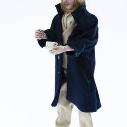 Miniature man in dark coat, light shirt, pants. Holds a cup and saucer. Has a hat and fair hair.