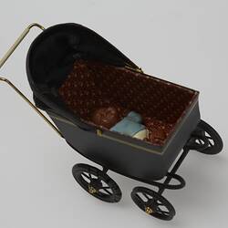 Black metal miniature pram. Contains baby doll with dark brown hair and skin wearing blue top and white pants.