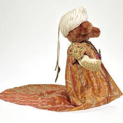 Profile of light brown plush bear wearing white turban, ornate tunic and coat with long tail.