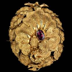 Gold oval locket brooch. Vine leaves and tendrils border. Branches at base with a small bird at top, all supporting a large flower with an amethyst purple paste stone centre.