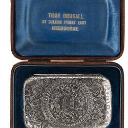 Back of heavily engraved silver cigarette case with central text. Rectangular with rounded corners. In blue lined case.