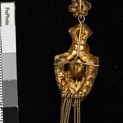 Back of gold pendant earring. Comprised of four connected sections with floral and leaf motifs. Gold fringe hangs from the bottom.