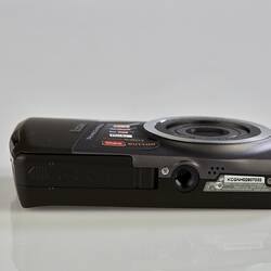 Black and grey camera laying flat, lens up. Base has screw in hole for tripod. Metal trim on sides and lens.