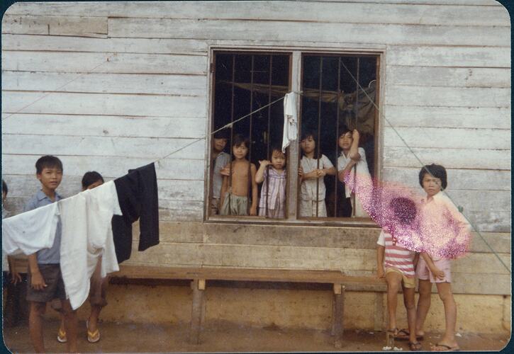 Children looking out the window of a white weatherboard building. Some stand either side of window. Clotheslines attache to window frame.