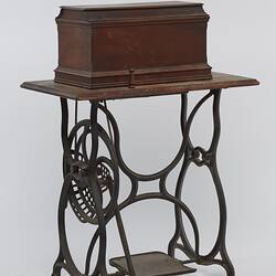 Sewing machine in wooden box fitted to a metal frame with a wooden bench and cover.