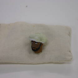 Small clay head of bearded man wearing a turban, lying on a cream cloth bag.