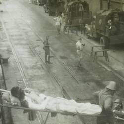 People unloading a stretcher with an injured person in bottom foreground, ambulances in background.
