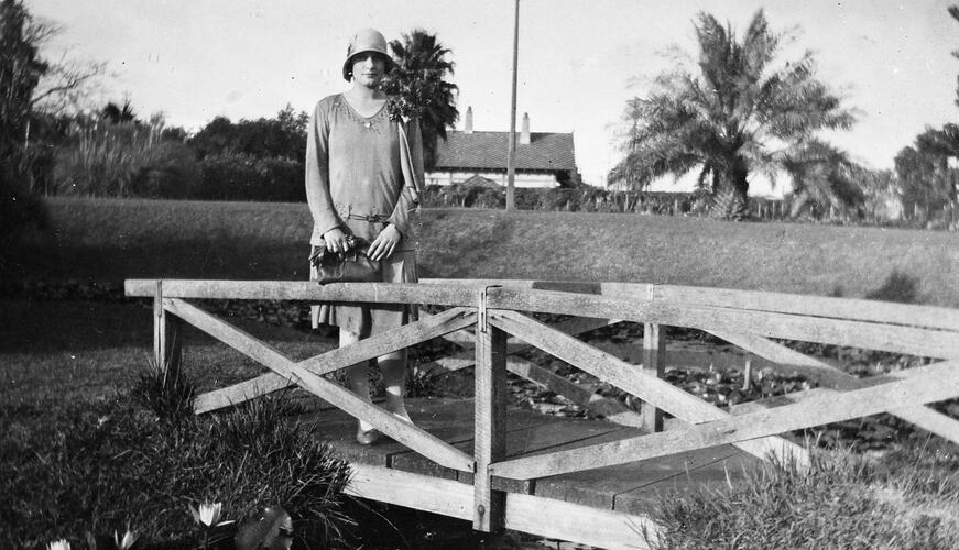 Woman wearing hat stands on wooden bridge in outdoor setting. Palm tree in background.