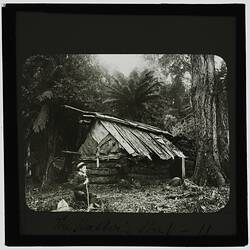 Black and white photograph of a boy standing in front of a rough wood hut.