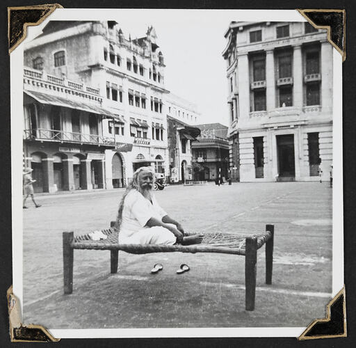 Man sitting on bed in street empty square with buildings in background.