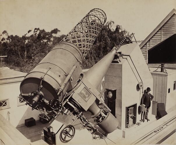 Great Melbourne Telescope with trees behind. Man standing at right wears suit and hat.