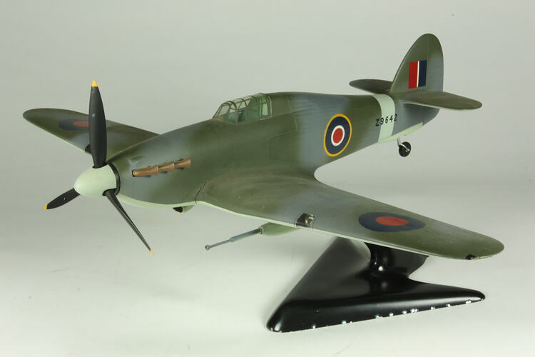 Green and grey model of single propeller aeroplane on black stand.