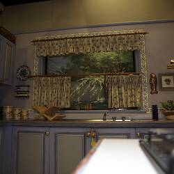 Kitchen studio set, pale blue cupboards below metal sink. Painted window above viewed from opposite stovetop.