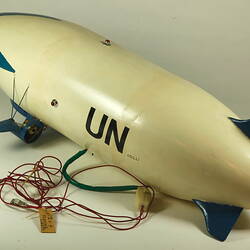 White model airship, blue nose and wings. Black UN on side. Small wheels, cables at base. Rear left view.