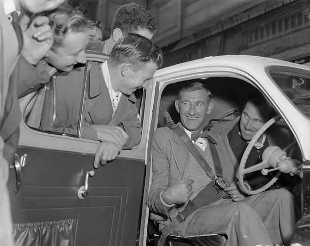 Two men sitting in car smiling at crowd looking in at them.