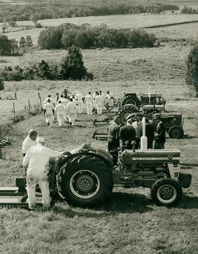 Men in white overalls walk past tractors in field.