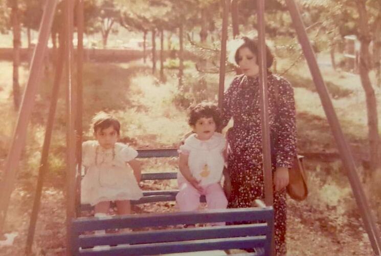 Woman standing beside two children seated on a swing in park.