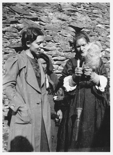 Young woman watches older woman with spindle of wool. Stone wall behind them.