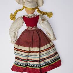 Back of doll with blonde plaits, white hat, wearing white shirt and red striped dress.