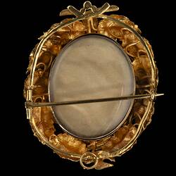 Back of gold oval locket brooch. Vine leaves and tendrils border. Branches at base with a small bird at top, all supporting a large flower with an amethyst purple paste stone centre.