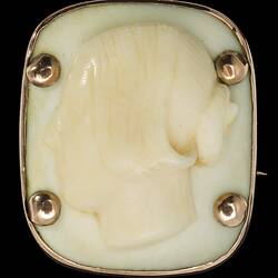 Square cameo with rounded corners featuring a white female bust facing left, hair up in a bun. Four prominent corner gold pins secure the cameo to its gold setting.