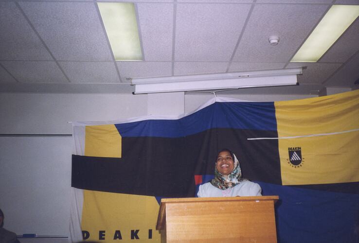 Woman standing at a lecturn delivering a speech. Blue, black and yellow flag behind her.