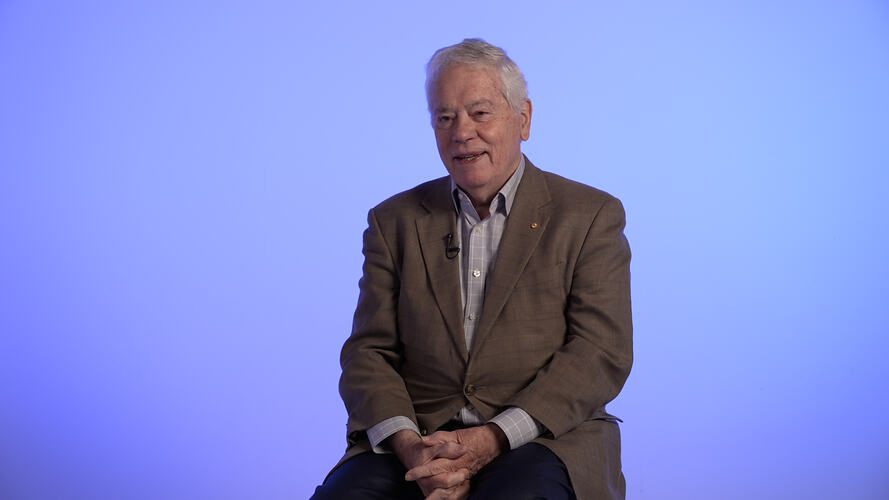 Seated man wearing brown jacket. Pale blue background.