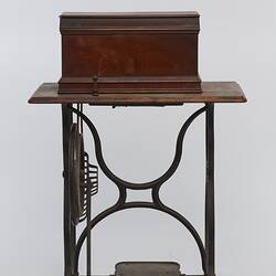 Sewing machine in wooden box fitted to a metal frame with a wooden bench and cover.