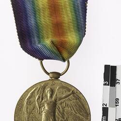 Round medal showing woman with wings holding plant leaf, with rainbow coloured ribbon.