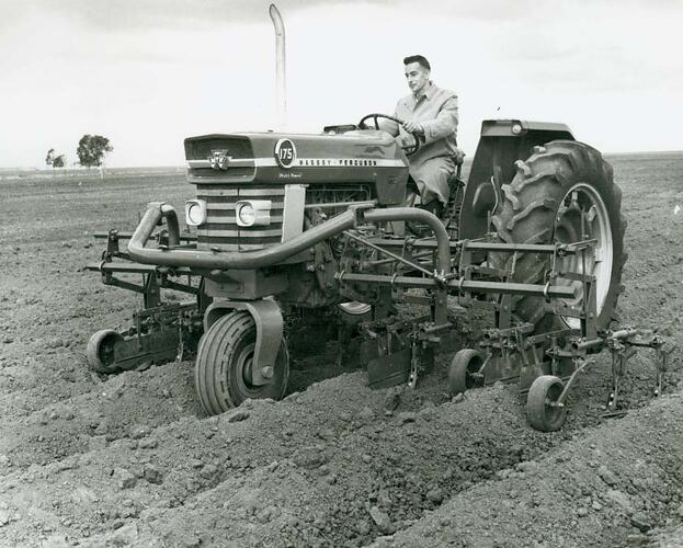 Front view of a man driving a tractor fitted with a mid-mounted cultivator across a wide open field.