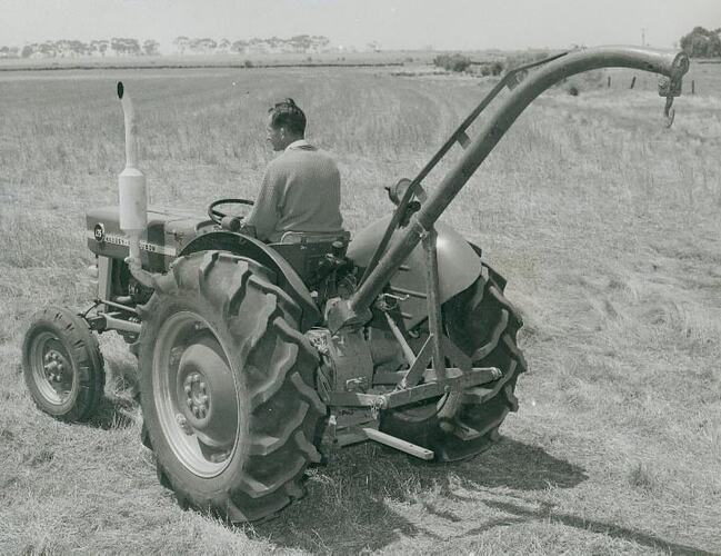 Rear view of a man driving a tractor with a jib crane attached across a field.