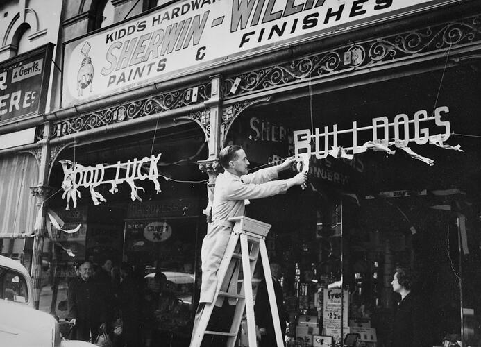 Man on a ladder hanging a sign outside a shop reading  'Good luck Bulldogs'.