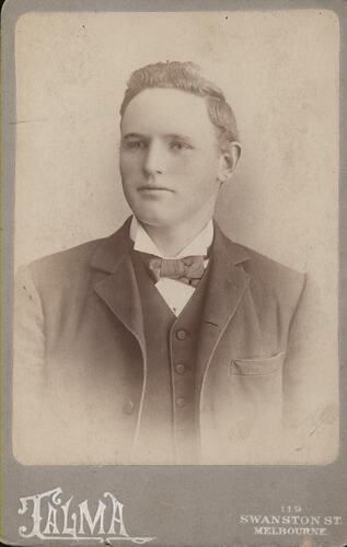 Male figure in formal pose wears a dark jacket and tie with a light shirt.