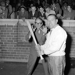 Man holding a baseball bat. Man behind has hands up as 'catcher'. Crowd behind them looks on.