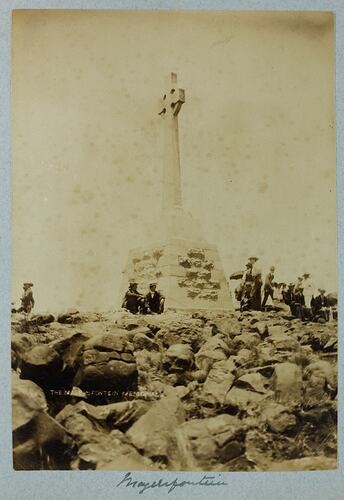 Group of men around a stone monument topped with a cross, rocky landscape in foreground.