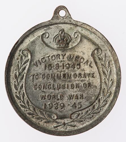 Round silver coloured medal with text on shield-shaped scroll framed by wreath. Loop at top.