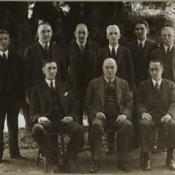 Formal group of nine men wearing suits in an outdoor setting. The three front men are seated.