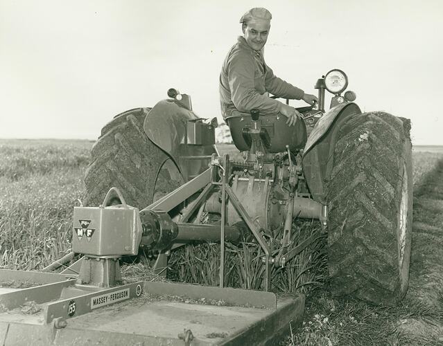 Man leaning over back of tractor seat, looking at a rotary slasher attached to back of tractor.