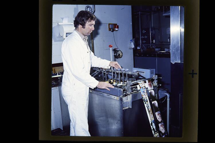 Man in white boiler suit tends to two horizontal reels of colour photographs.
