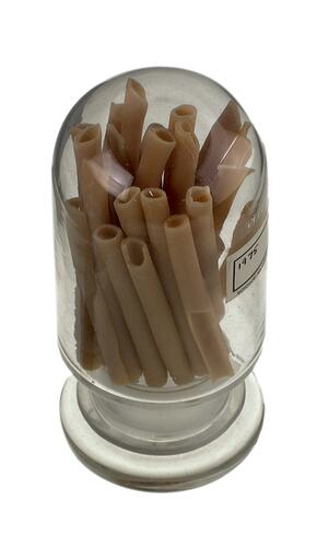 Clear glass jar containing brown long tube-shaped pasta. Three quarter view from above.