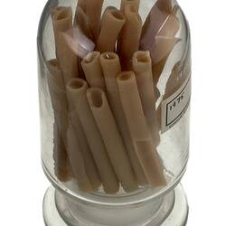 Clear glass jar containing brown long tube-shaped pasta. Three quarter view from above.