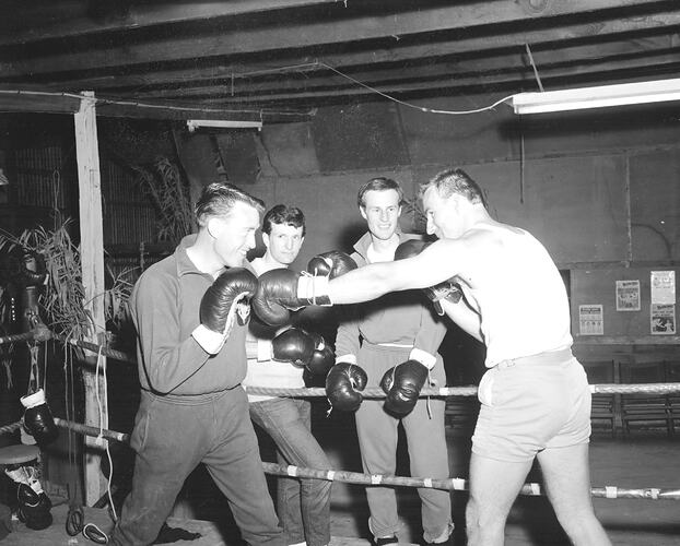Two men watching two other men sparring in a boxing ring.