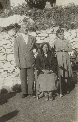 Father and daughter stand either side of seated, veiled mother. Stone wall behind them.