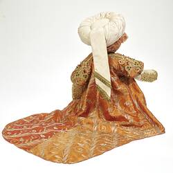Back view of teddy bear wearing white turban, ornate embroidered coat with long tail.
