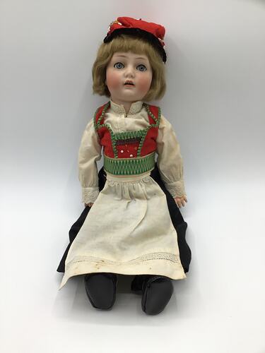 Seated doll with red, green, white top. White apron on black skirt. Red hat. Brown hair, blue eyes.