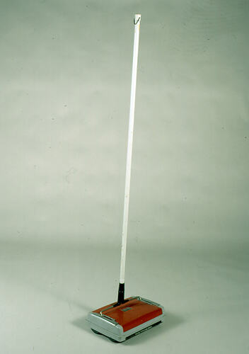 Carpet sweeper, white handle with red and silver sweeper pan, shown upright.