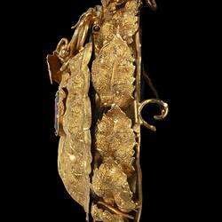 Profile of gold oval locket brooch. Vine leaves and tendrils border. Branches at base with a small bird at top, all supporting a large flower with an amethyst purple paste stone centre.