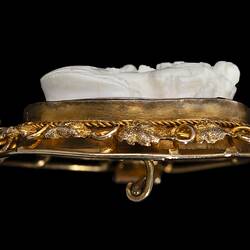 Profile of oval brooch with gold framing white cameo relief woman facing right. Frame is gold leaves and tendrils.