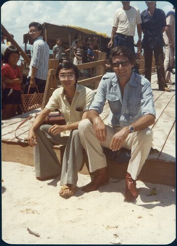 Two men sit on edge of wooden walkway, their feet on the sand. Crowd behind them.