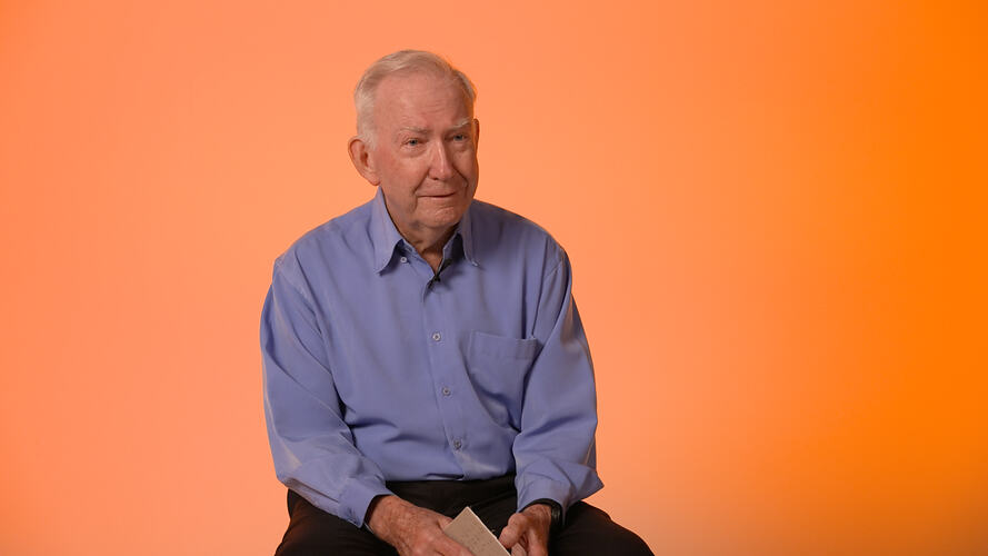 Seated man wearing blue shirt with orange background.